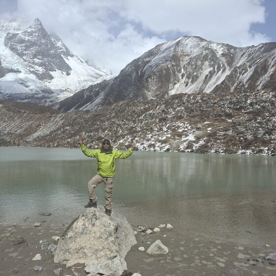 Navaraj Thapa standing on a boulder at a glacial lake in Langtang, snow peaks behind