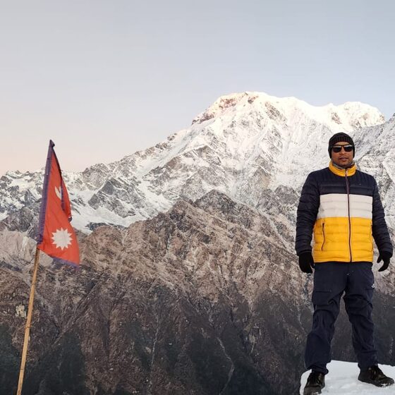 Navaraj Thapa on a ridge at Mardi Himal at dawn, a Nepali flag beside him and Annapurna South behind